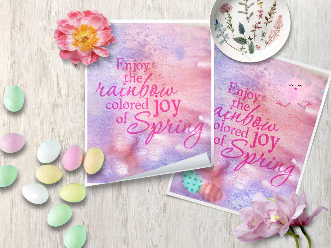 Easter themed pink and purple art prints laying on wooden surface with easter eggs, flowered plate, and flowers scattered around
