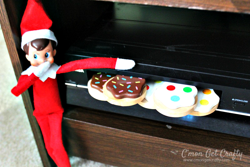 Elf on the Shelf sitting with hand over mouth in front of a VCR with toy cookies sticking out of the tape slot