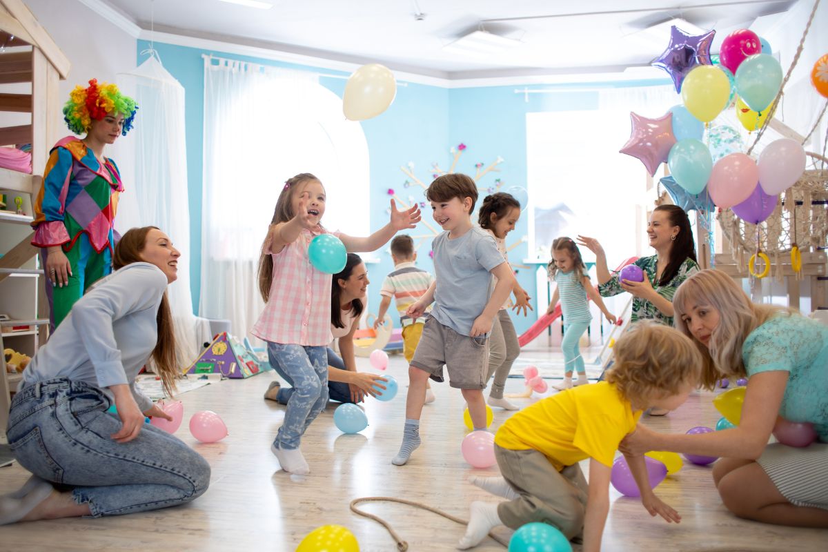 Party scene with mix of adults and children, balloons in light room with pale blue walls and bright windows