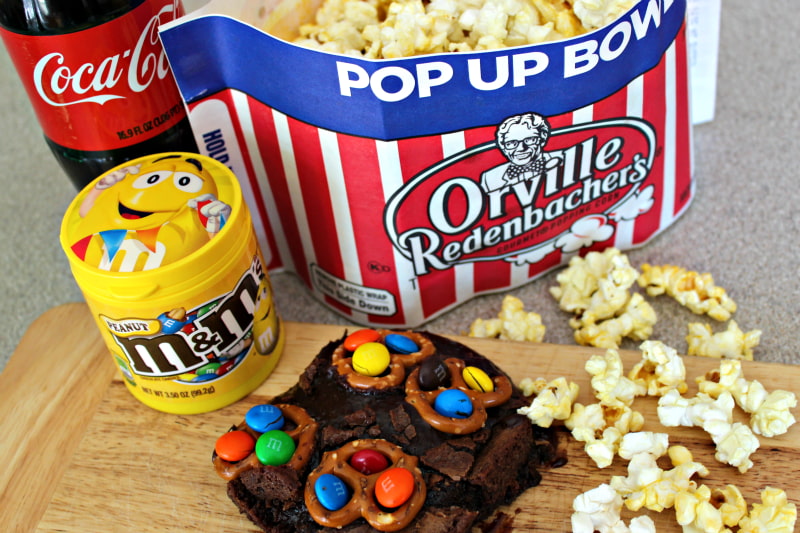 Popcorn in a bag and also spilled on counter over wooden board next to brownie topped with pretzels and M&Ms, a yellow M&M container, and a plastic coke bottle.