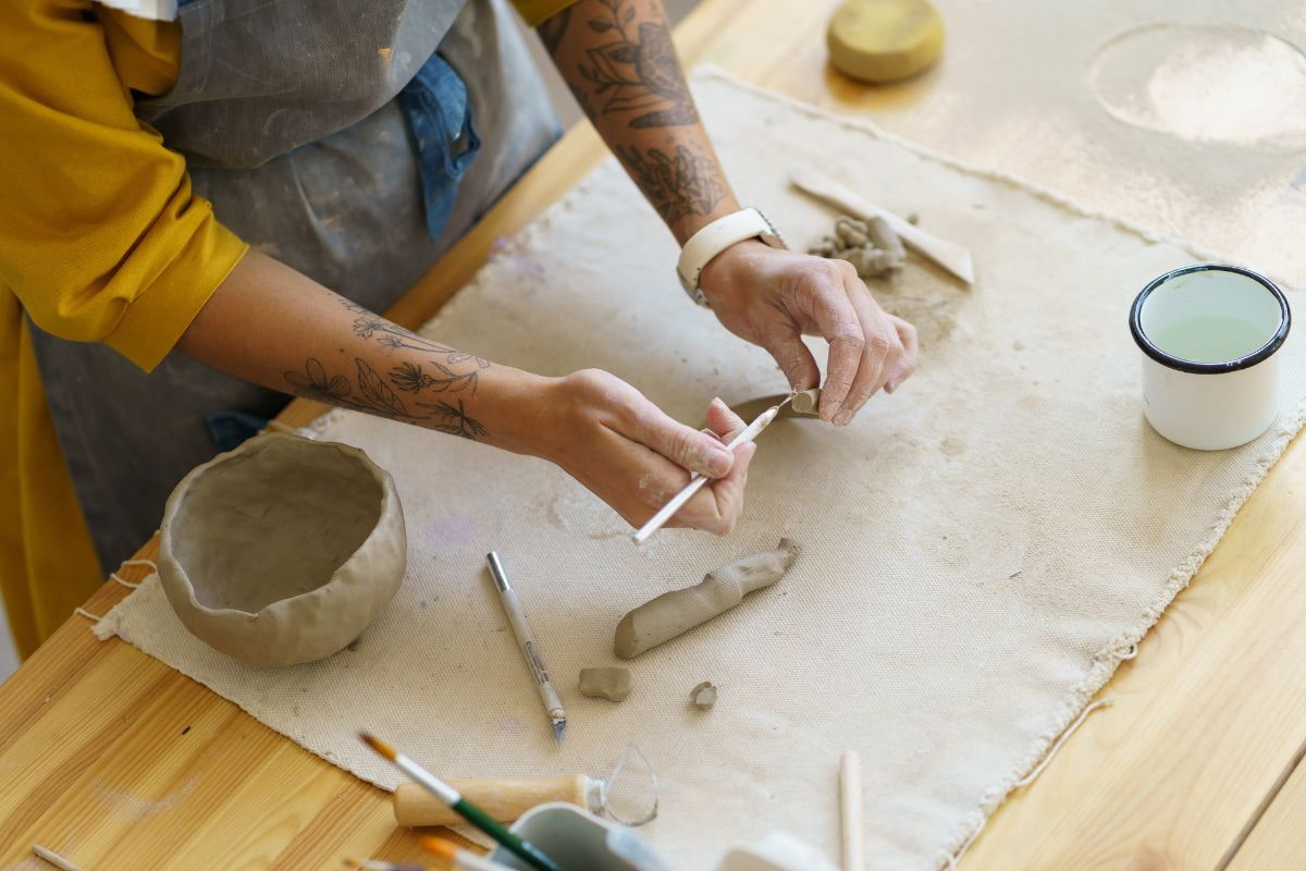 hands working clay tools on clay with ceramic mat, clay, and partically created clay bowl laying nearby.