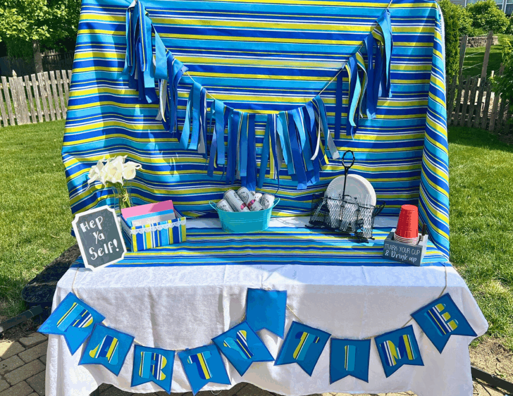 1 - C'mon Get Crafty blue and green colored party table set up with rag banner, pennant banner, fabric backdrop, white tablecloth, and table runner. table set with picnic and outdoor dining decor - landscape style image