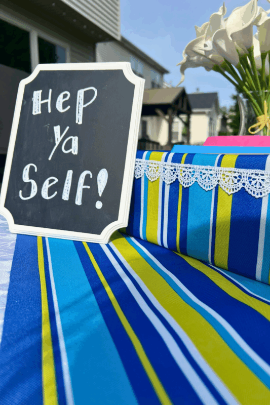 7 - C'mon Get Crafty close-up image of chalkboard sign "Hep Ya Self" sitting on table next to fabric covered box with striped table runner and white flowers in a vase