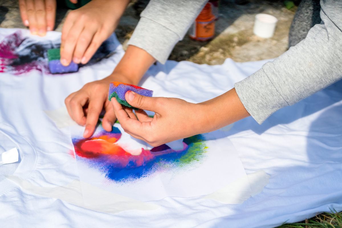 EyeCandyCustomz-387879-Fabric-Paint-Shirt-image1 1 - C'mon Get Crafty children's hands applying multicolored paints to white t-shirt with sponges