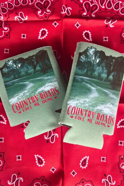 2024 Farm Party Portrait pic edits - C'mon Get Crafty Two green can coolers with a picture of a country landscape on them. The text below the image says “Country Roads Take Me Home” in red. They are lying on a red bandana.