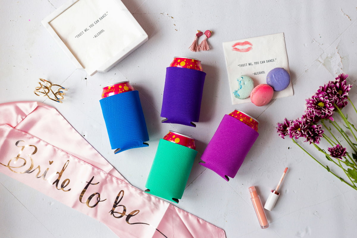 A view of a flatlay featuring four cans, each dressed in a different color can cooler (blue, purple, green, and pink). The flatlay also includes accessories such as a pink "Bride to be" sash, flowers, and macarons.
