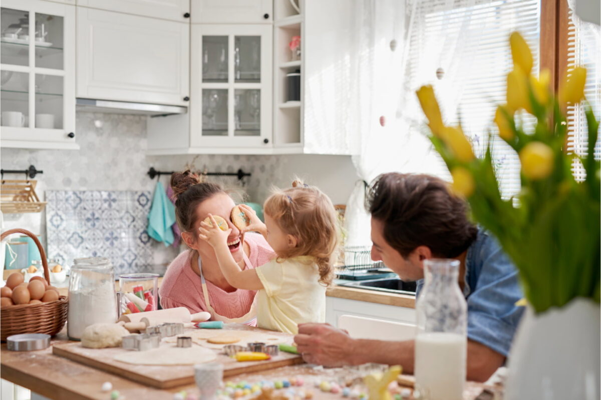 A joyful moment in a bright kitchen where a young girl playfully holds decorated cookies over her mother's eyes while the father laughs nearby. The countertop is covered with baking ingredients, cookie cutters, and colorful decorations, suggesting a fun family baking session. Yellow tulips in the foreground add a cheerful touch to the cozy scene.