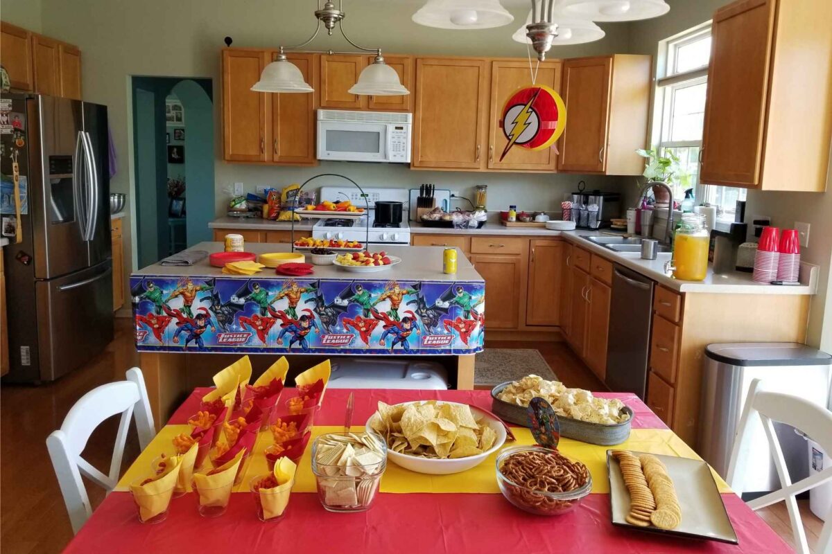 Henrys 5th Birthday Party - C'mon Get Crafty A kitchen decorated for a superhero-themed party, featuring Justice League table coverings and a Flash symbol hanging from the ceiling. The counters and table are filled with snacks like chips, pretzels, crackers, cheese puffs, fruit skewers, and a large drink dispenser filled with orange juice. Red and yellow colors dominate the décor, including paper cups, napkins, and snack containers.