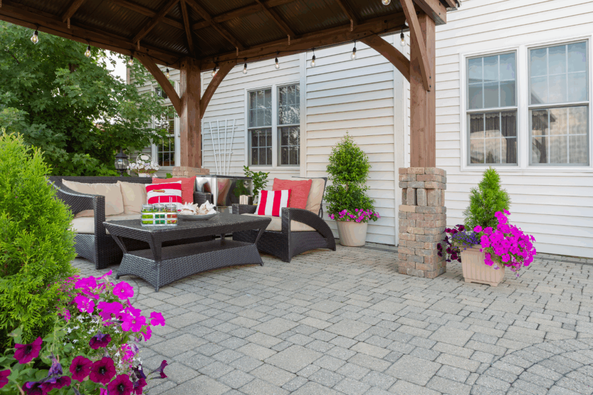 Patio Picture - C'mon Get Crafty A cozy backyard patio setup under a wooden pergola with string lights, featuring wicker furniture with beige cushions and red-striped pillows. The space is surrounded by potted greenery and vibrant purple and pink flowers, creating a welcoming and colorful outdoor retreat. A coffee table holds drinks and snacks, adding to the relaxed atmosphere.